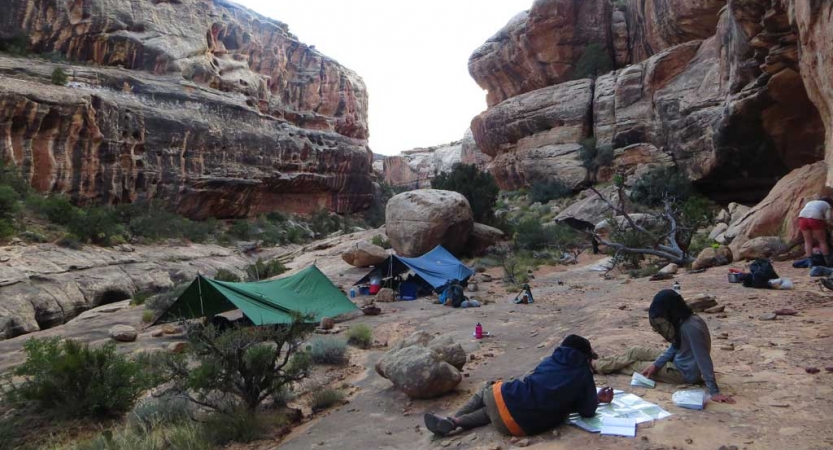 Tarp shelters and a few people rest at a campsite nestled between canyon walls. 